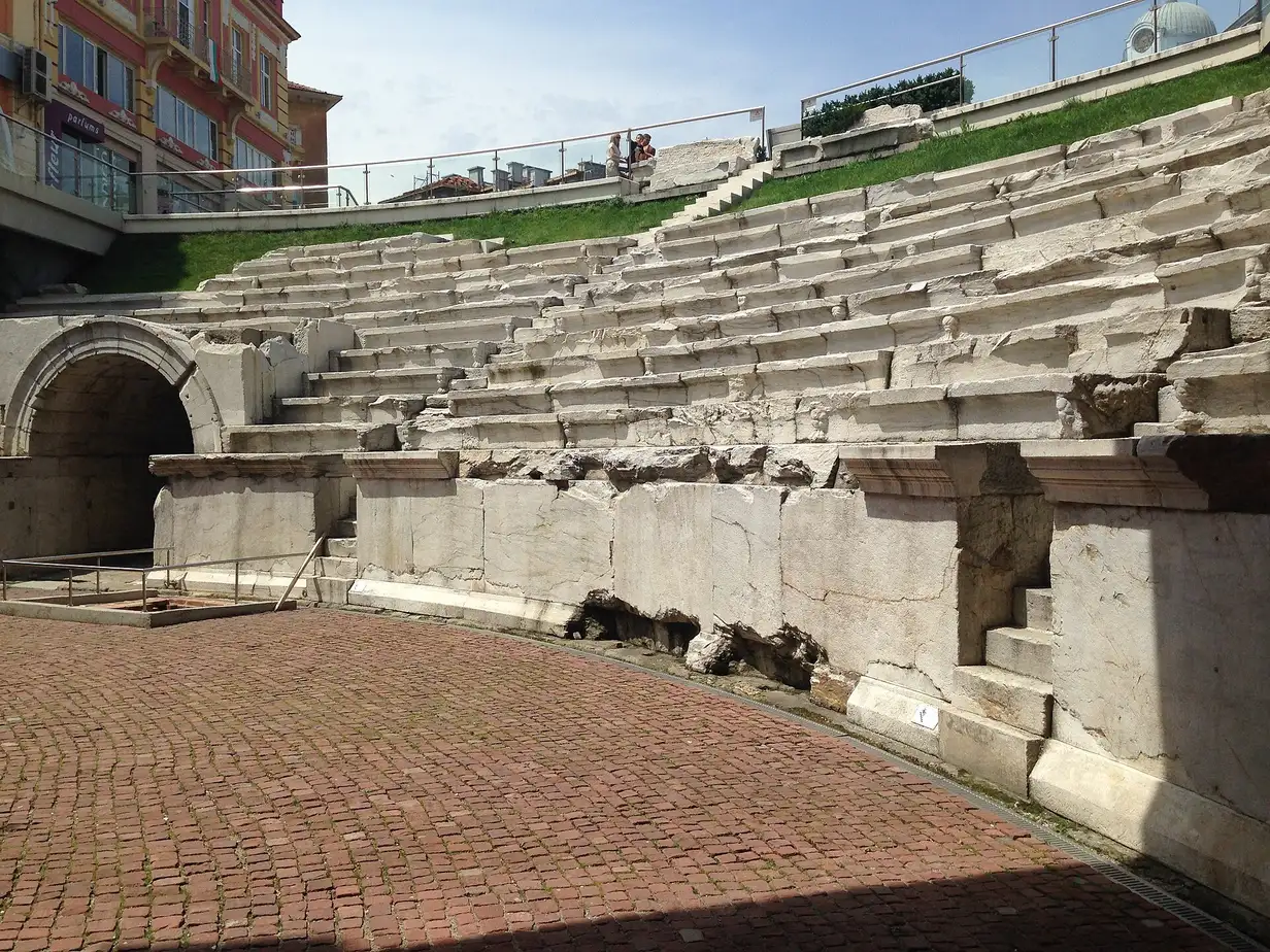 The curved marble seating rows of the Roman Stadium of Philippopolis sunk below street level at Dzhumaya Square, with a vaulted entrance arch and modern buildings above