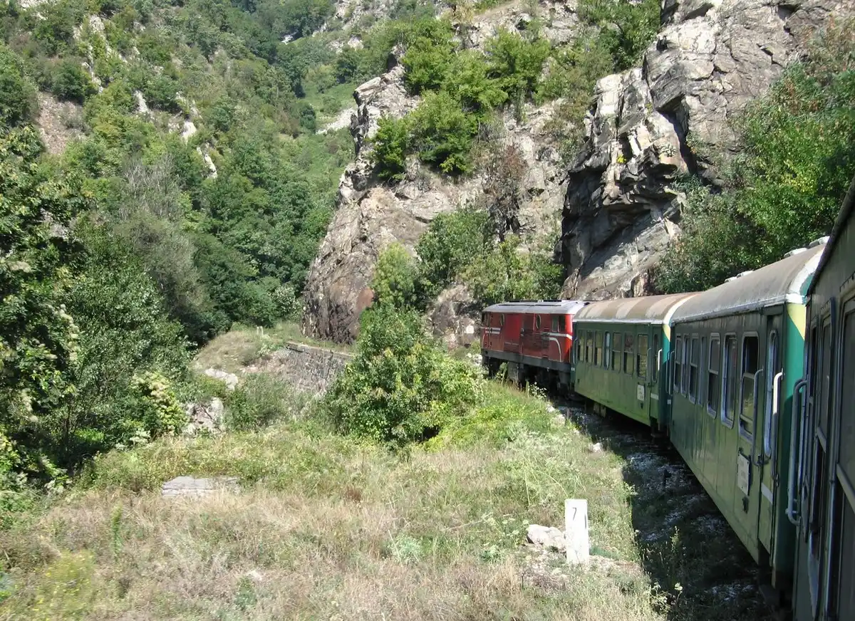 A red narrow-gauge diesel locomotive hauling green carriages through a rocky Rhodope gorge