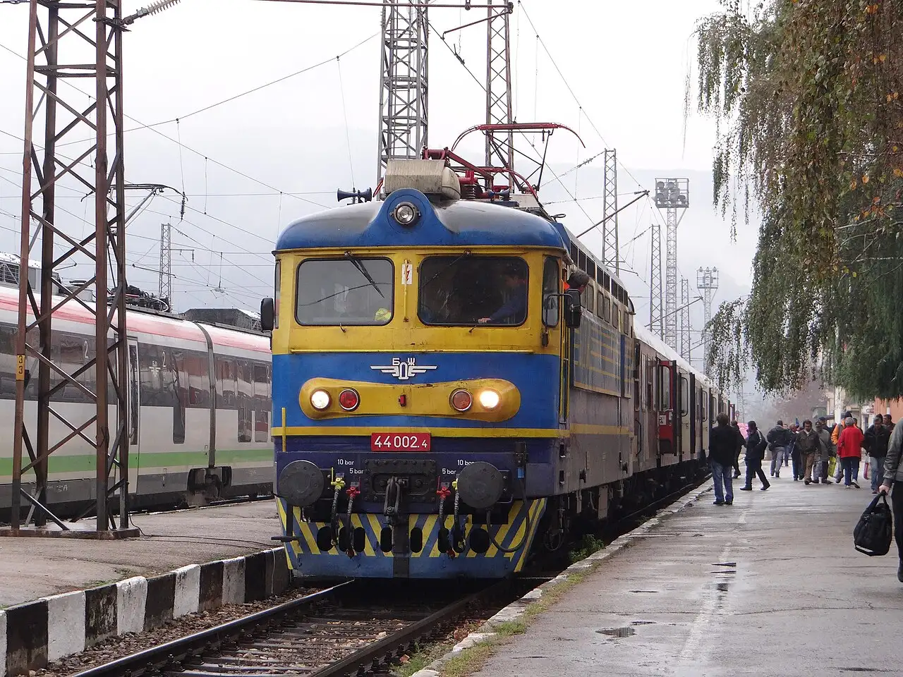 A blue and yellow Bulgarian State Railways locomotive at a platform with passengers waiting