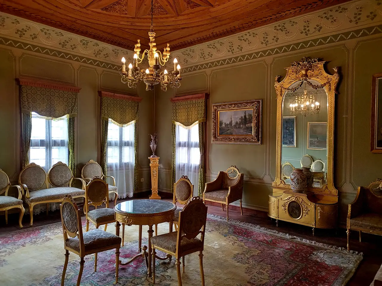 A Revival-era salon with a carved wooden ceiling, a brass chandelier, gilded chairs around a small round table and a tall gilt mirror