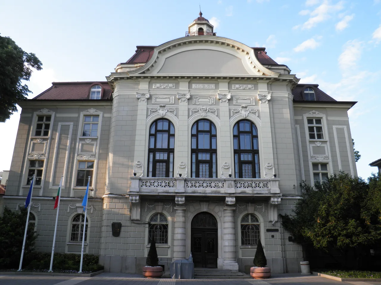 The cream neoclassical facade of Plovdiv's municipality building, with Baroque detailing, three tall arched windows over a stone balcony and flags out front