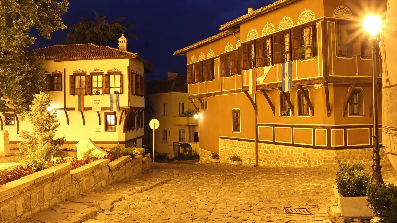 A cobbled lane in Plovdiv Old Town at dusk, lamplight on ochre and white Revival houses with carved eaves and wooden shutters