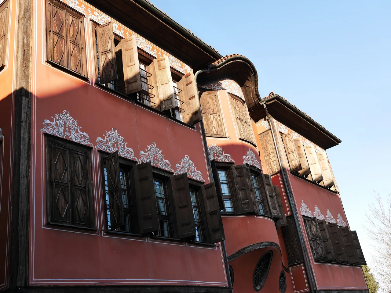 The red facade of the Georgiadi House in Plovdiv Old Town, with white painted ornament, rows of wooden shutters and a curved central bay