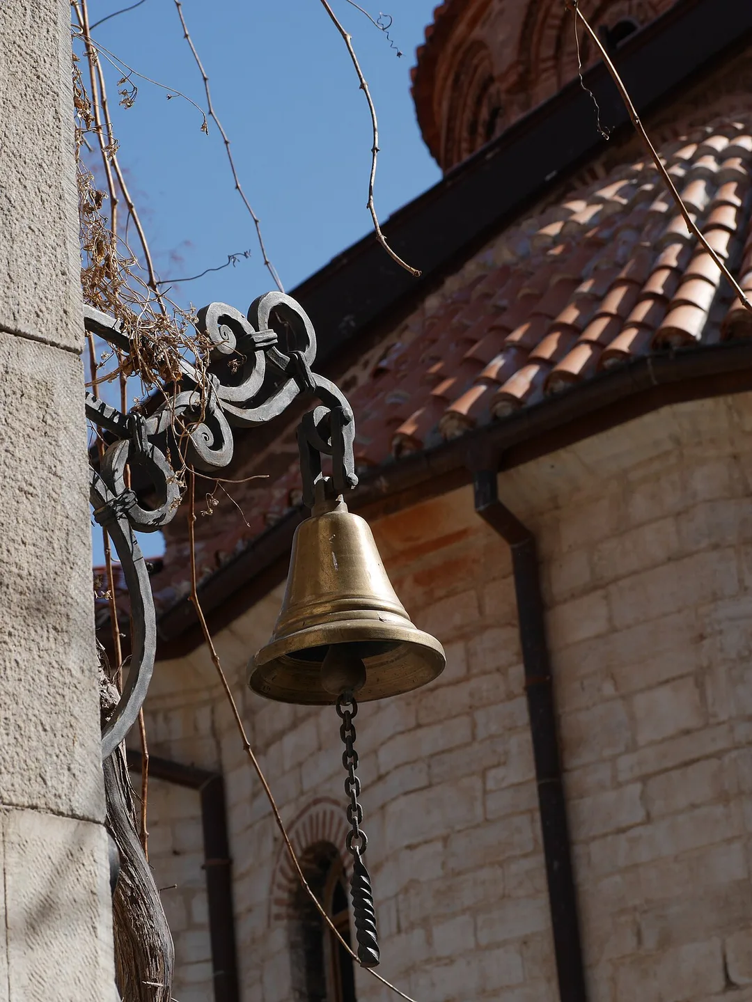 A bronze bell on a wrought-iron bracket against the tiled dome and stone wall of Bachkovo Monastery church