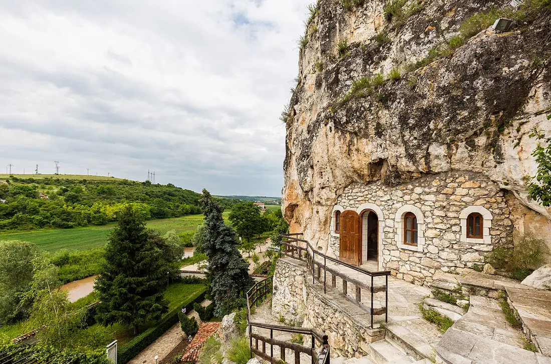 The Basarbovo Rock Monastery near Ruse, a stone facade with arched doors and windows built into a limestone cliff above a green valley, reached by stone steps