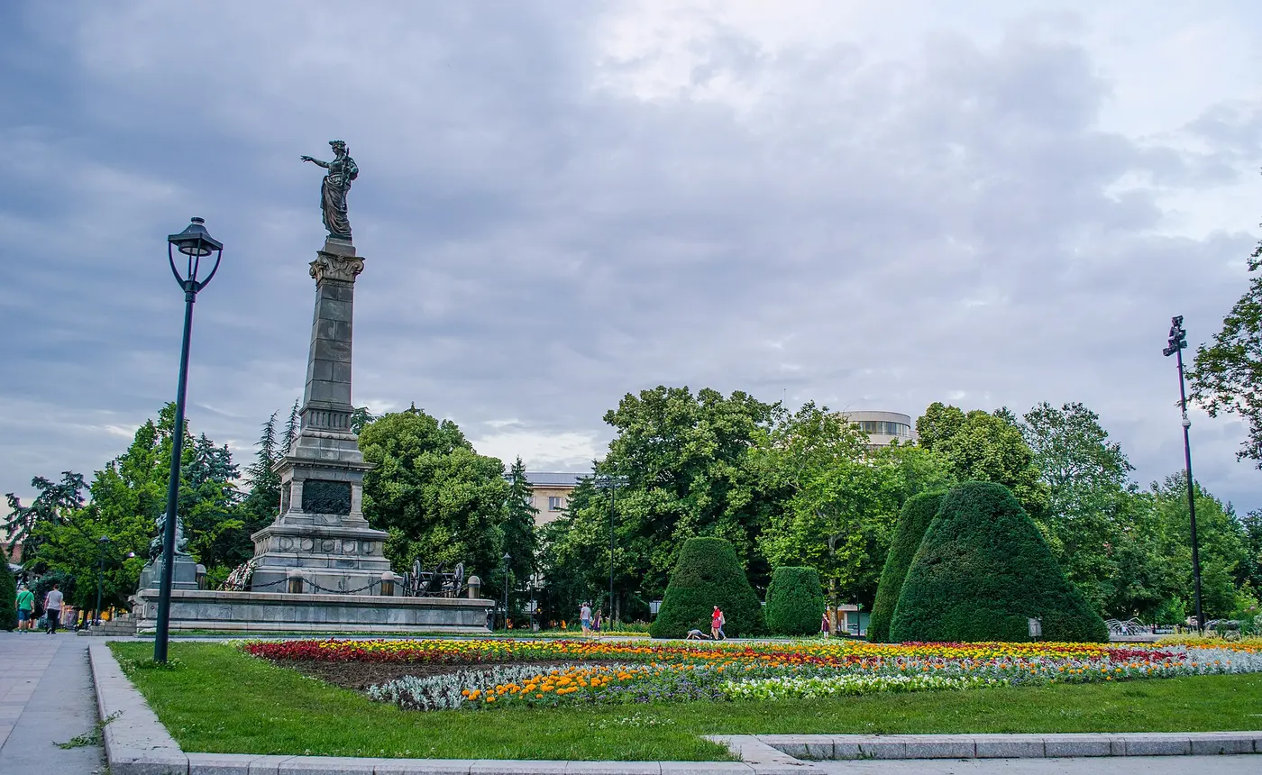 The Monument of Liberty on Freedom Square in Ruse, a tall stone column topped by an allegorical female figure, with bright flowerbeds and clipped topiary around it