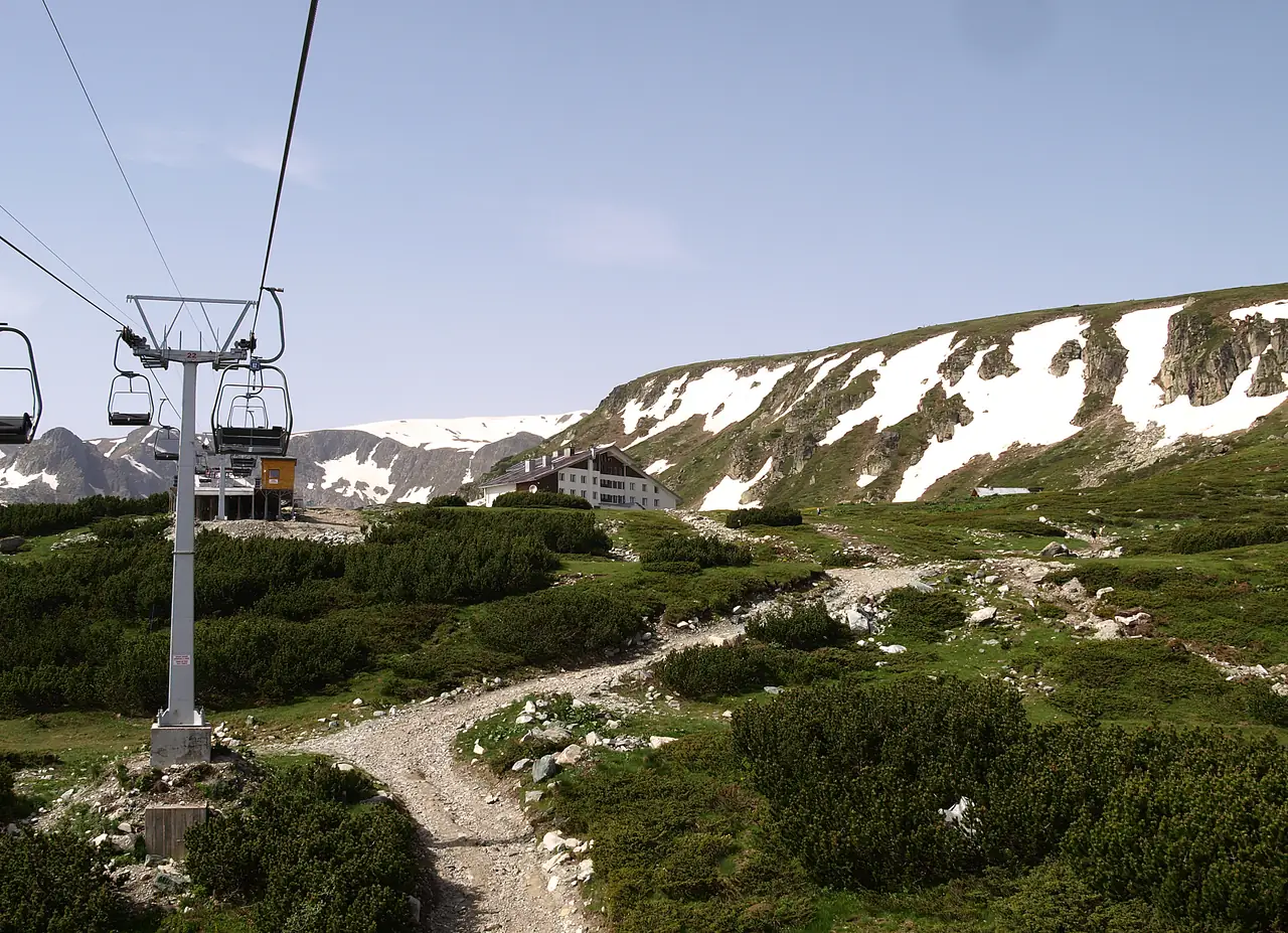 A chairlift climbing over a rocky trail toward the Rila Lakes hut, with snow patches on the ridge behind