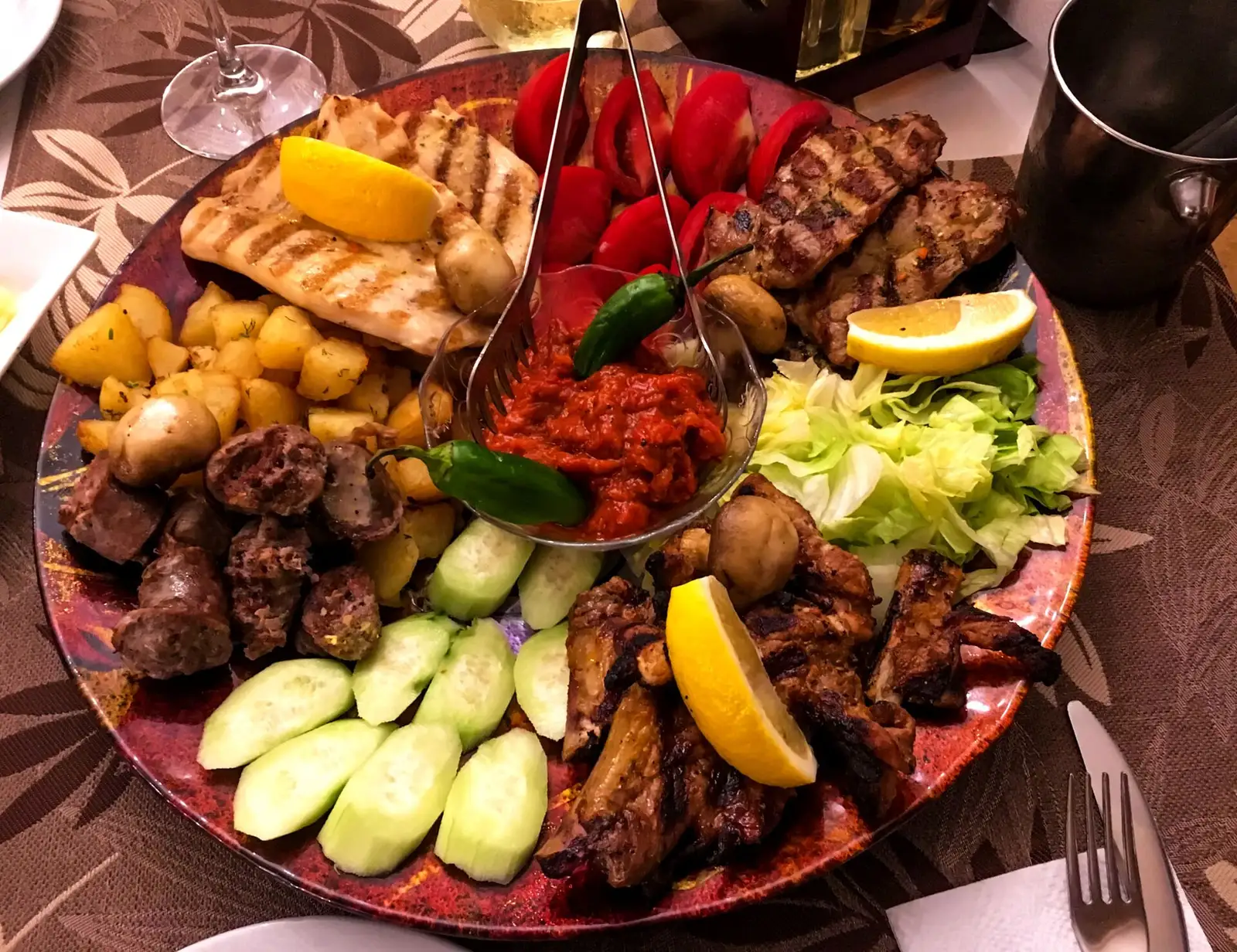 A large mixed grill platter of Bulgarian meats with roasted potatoes, tomatoes, cucumbers and a bowl of lyutenitsa pepper sauce