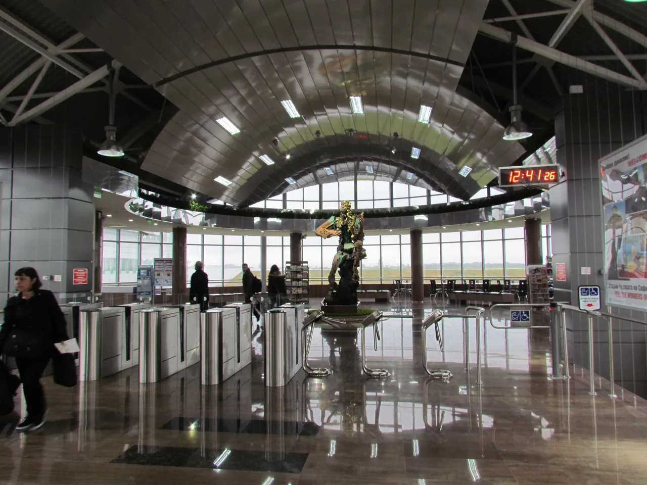 The concourse of Sofia Airport metro station with ticket gates, ticket machines, a statue and a departures clock