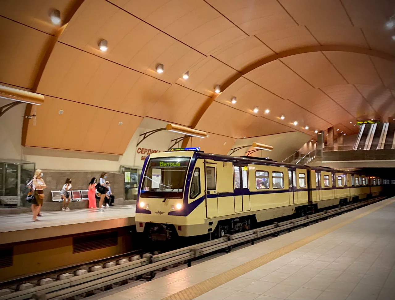 A cream and blue Sofia metro train at Serdika station in the centre, passengers waiting under a curved ceiling