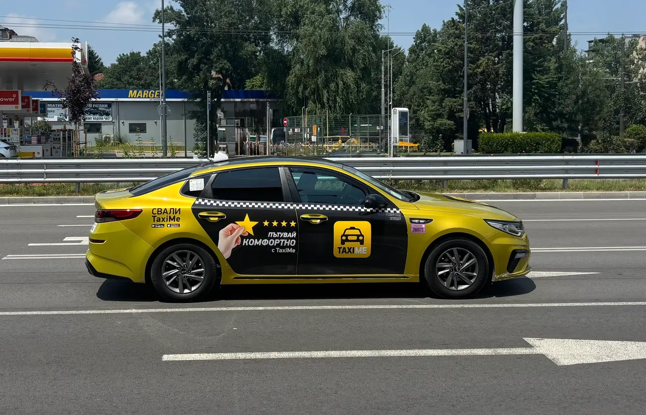 A bright yellow Sofia taxi with TaxiMe branding driving along a road with a petrol station behind