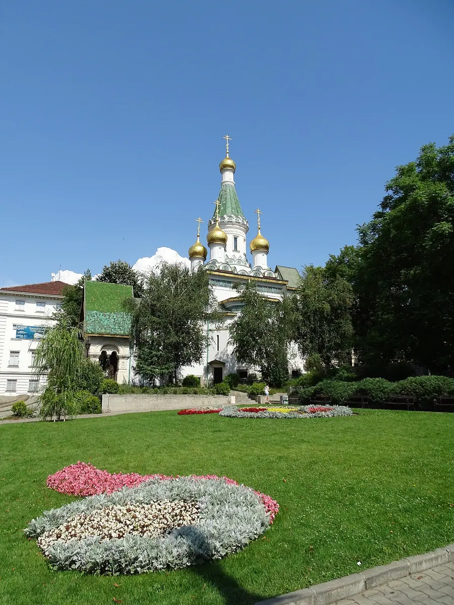 The golden onion domes and majolica-tiled facade of the Russian Church of St Nicholas in Sofia above a flower garden