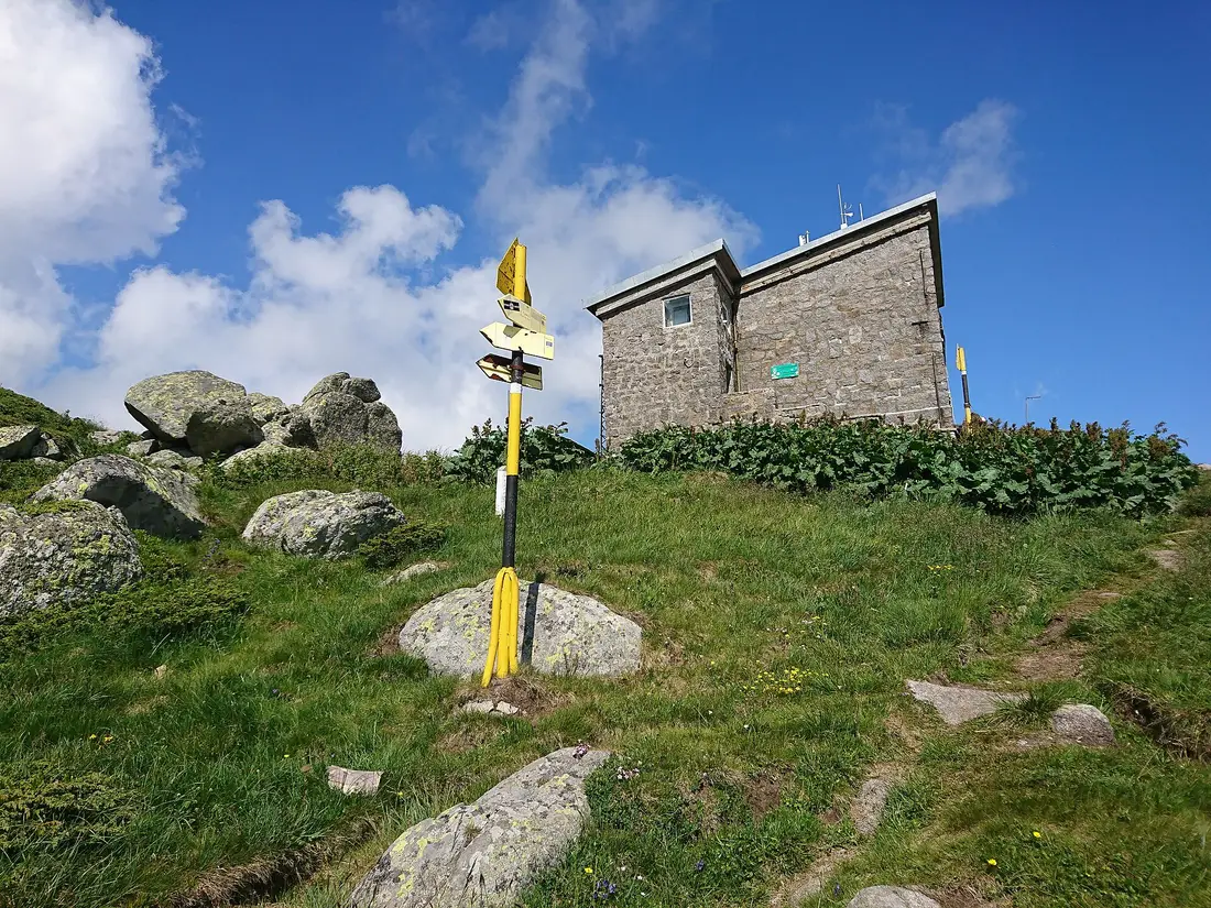 A yellow trail marker post and the stone weather station building on the bare summit plateau of Cherni Vrah on Vitosha