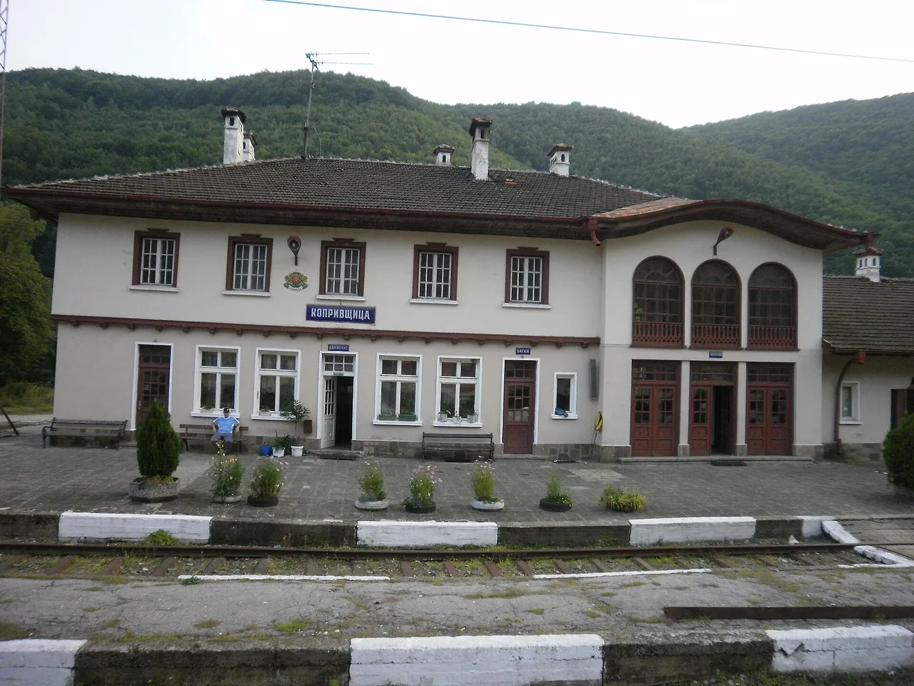 The small white and red Koprivshtitsa railway station building with forested hills behind and empty platform tracks in front