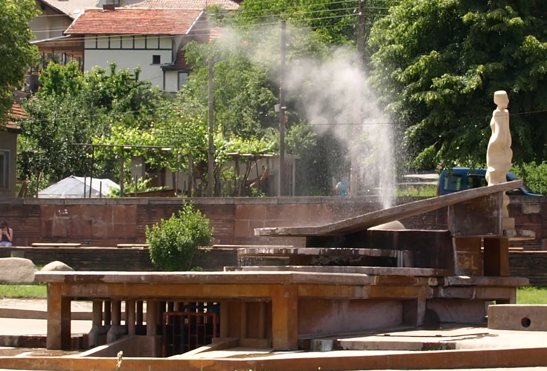A jet of steaming water shooting up from the stone geyser basin in the centre of Sapareva Banya with village houses behind