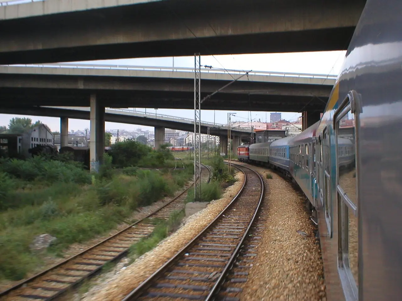 A passenger train curving on the tracks into Belgrade beneath road flyovers with the city skyline behind