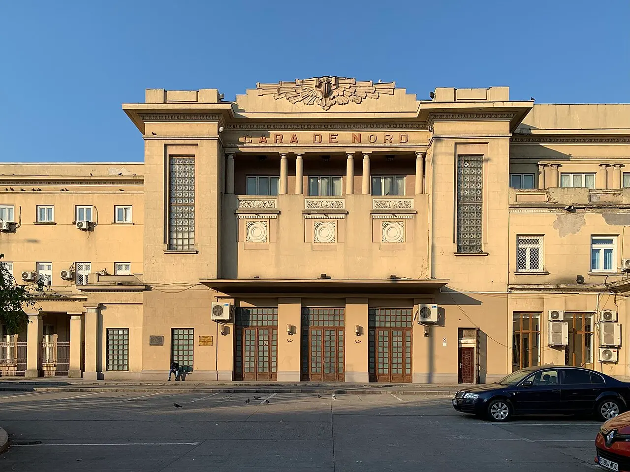 The tall inter-war stone facade of Bucharest Gara de Nord station at golden hour, with the carved GARA DE NORD lettering and a car parked in front