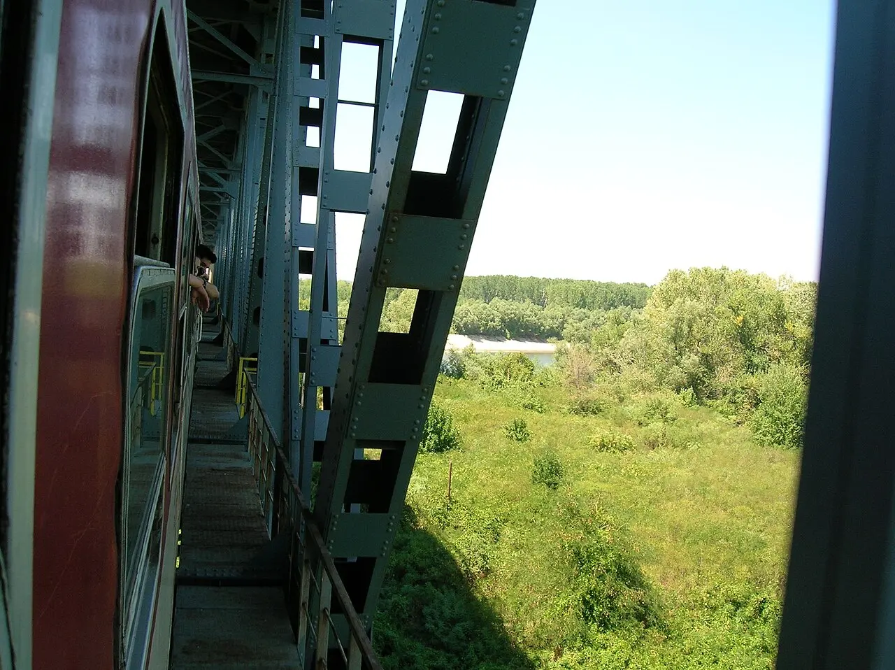 View from a train window along the green steel girders of the Danube Friendship Bridge, with the river and wooded banks below
