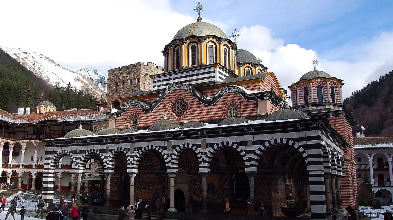 The striped arches and domes of Rila Monastery church with snow-dusted mountains rising behind the walls