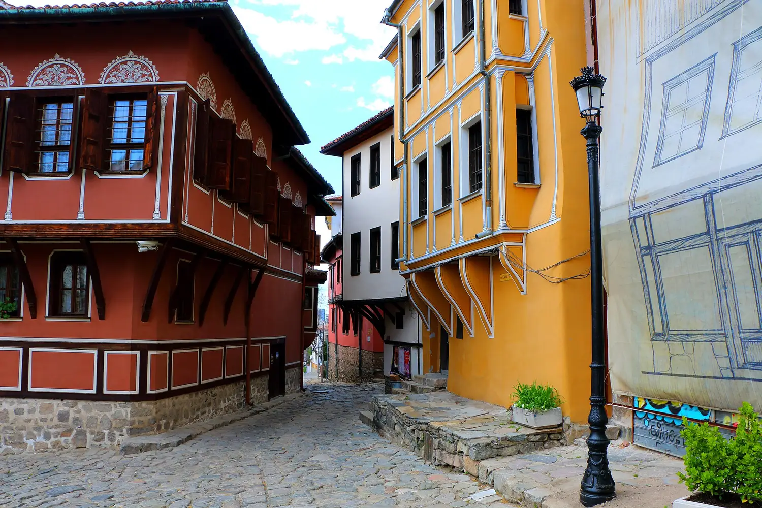 A cobbled lane in Plovdiv Old Town between a deep red and a yellow National Revival house with overhanging upper floors and a black street lamp