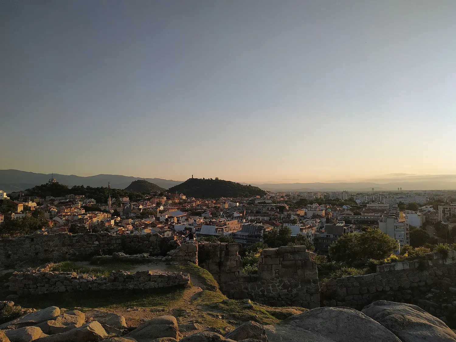 Plovdiv at sunset seen from the Thracian ruins of Nebet Tepe, city rooftops below and hills with a monument silhouette against a golden sky