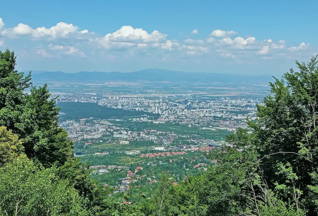 Panorama of Sofia spreading across the plain seen from the forested slopes of Mount Vitosha, distant mountains on the horizon