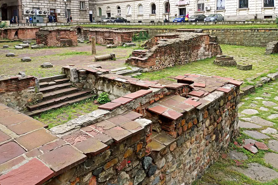 Exposed brick and stone ruins of ancient Roman Serdica in central Sofia, with modern buildings and parked cars behind