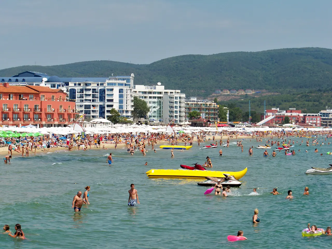The wide sandy beach at Sunny Beach with swimmers and a banana boat, hotels and green hills behind