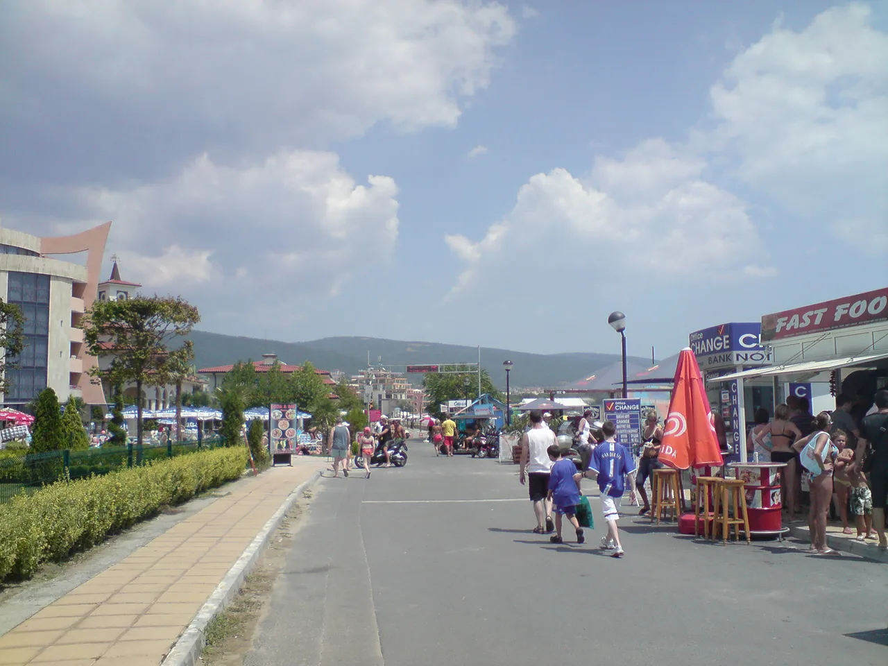 The main pedestrian strip at Sunny Beach lined with bars, a currency exchange and a club sign, running towards the hills