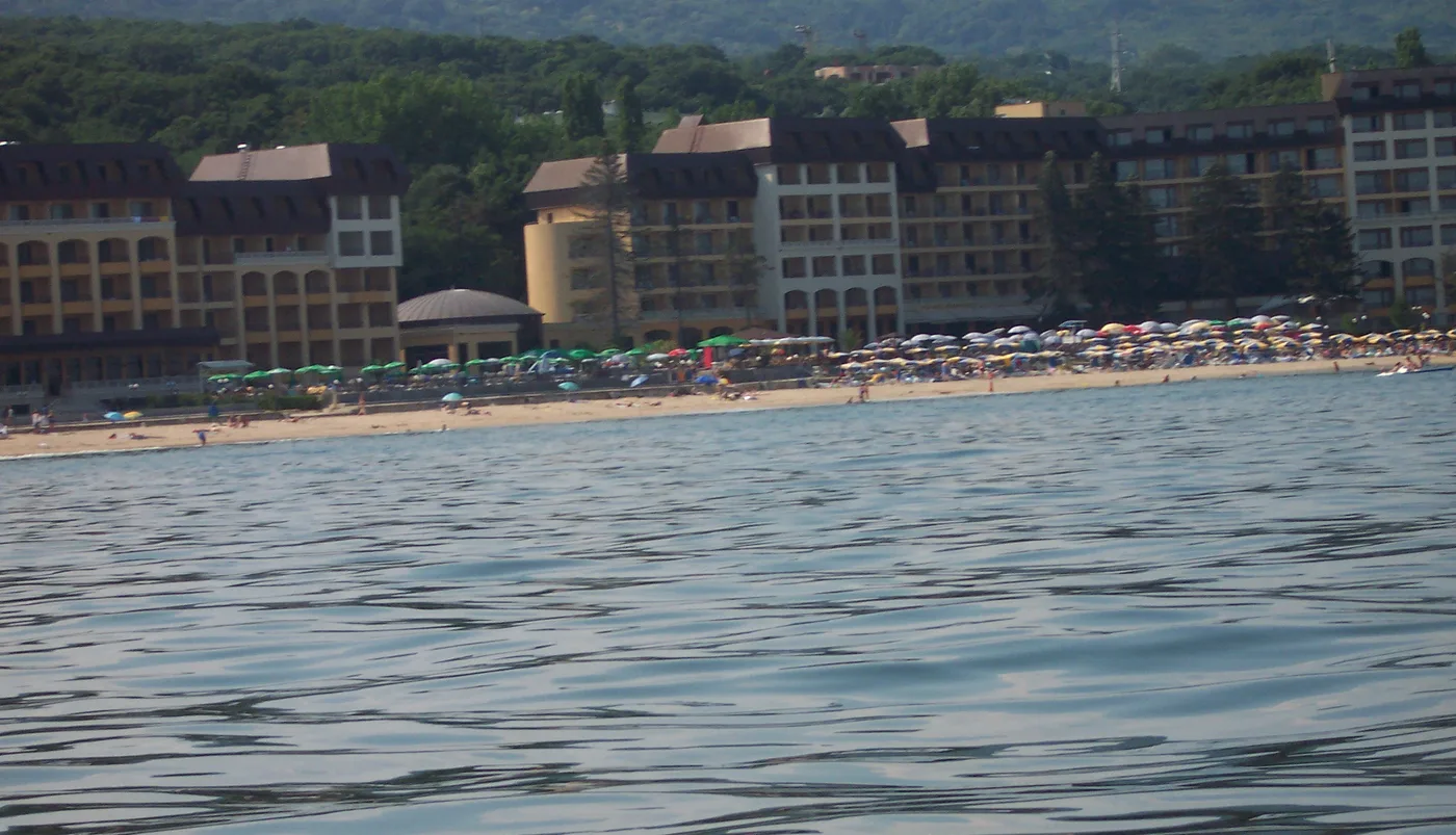 The beach at Golden Sands seen from the water, rows of umbrellas and low resort hotels with brown roofs backed by the green forested hills of the nature park