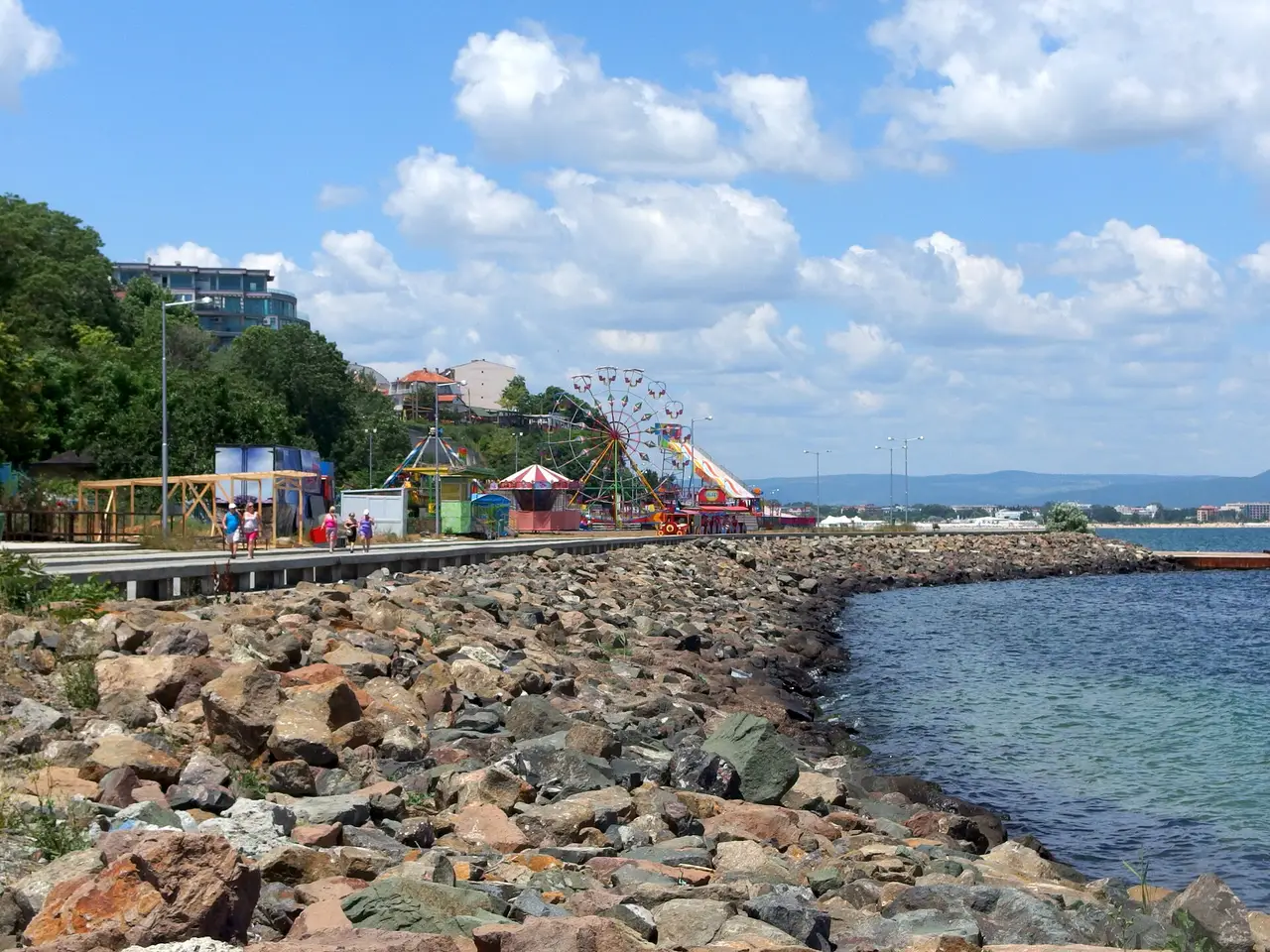 The seafront promenade at Nessebar with a small funfair and Ferris wheel, a rock breakwater curving into the bay and the resort coast stretching into the distance