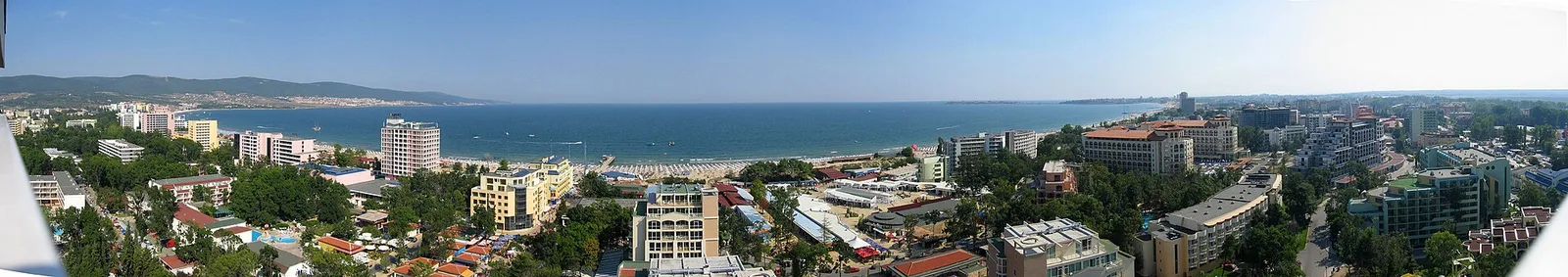 Aerial panorama of Sunny Beach: the long curve of the bay lined with a dense wall of hotels and a wide sandy beach, green hills rising behind the resort