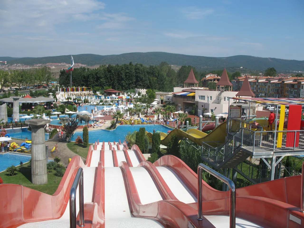 View from the top of a red water slide at Action Aquapark in Sunny Beach, looking over pools, a mock castle and the resort with green hills behind