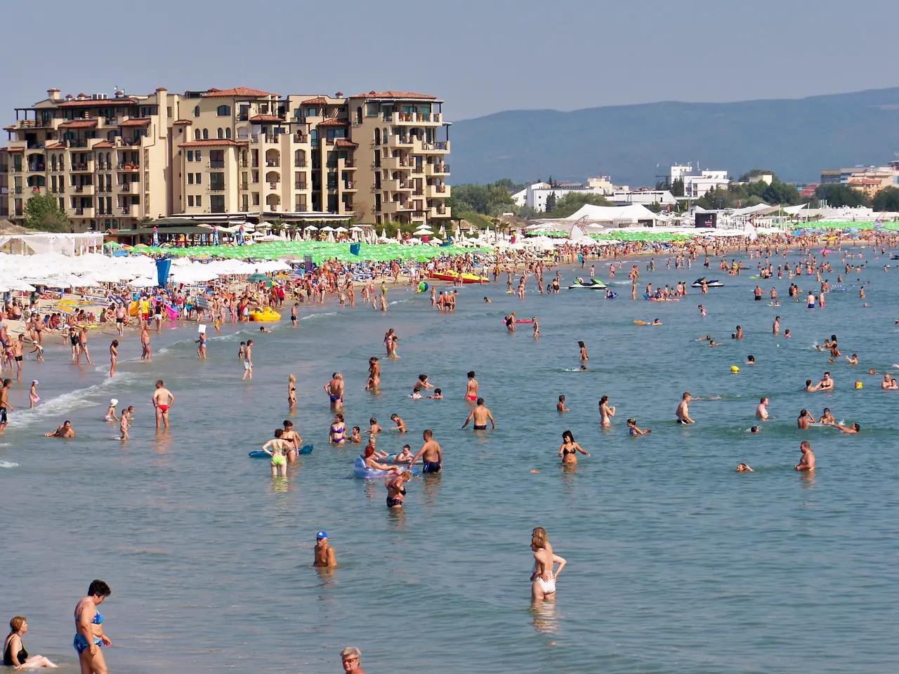 The packed sandy beach of Sunny Beach in high summer, crowds in the shallow water with a large hotel block and green hills behind