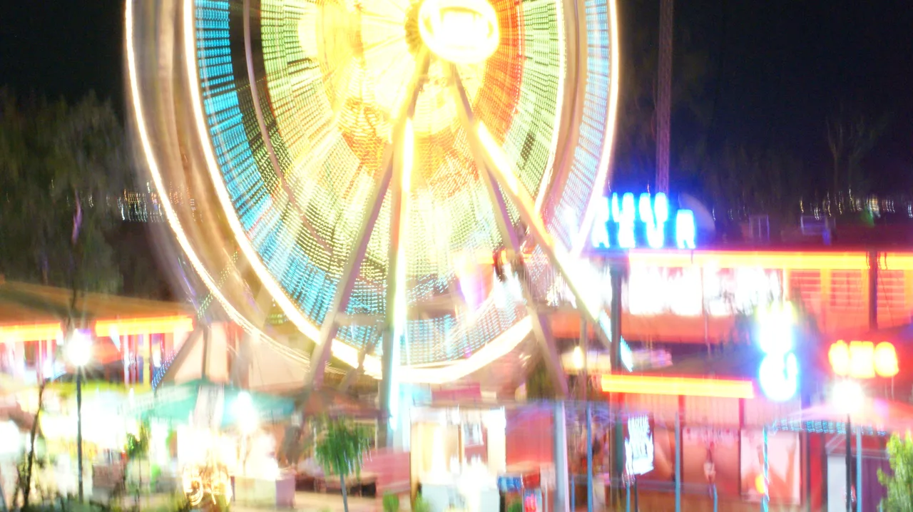 A brightly lit fairground Ferris wheel spinning at night at Sunny Beach, its lights blurred into streaks, with neon-signed bars beside it