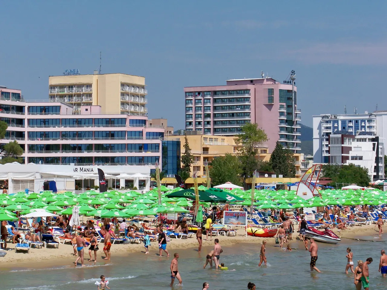 Green beach parasols and sunbeds packed along the Sunny Beach shore in front of a row of pastel high-rise hotels, with a jet-ski rental and a beach bar