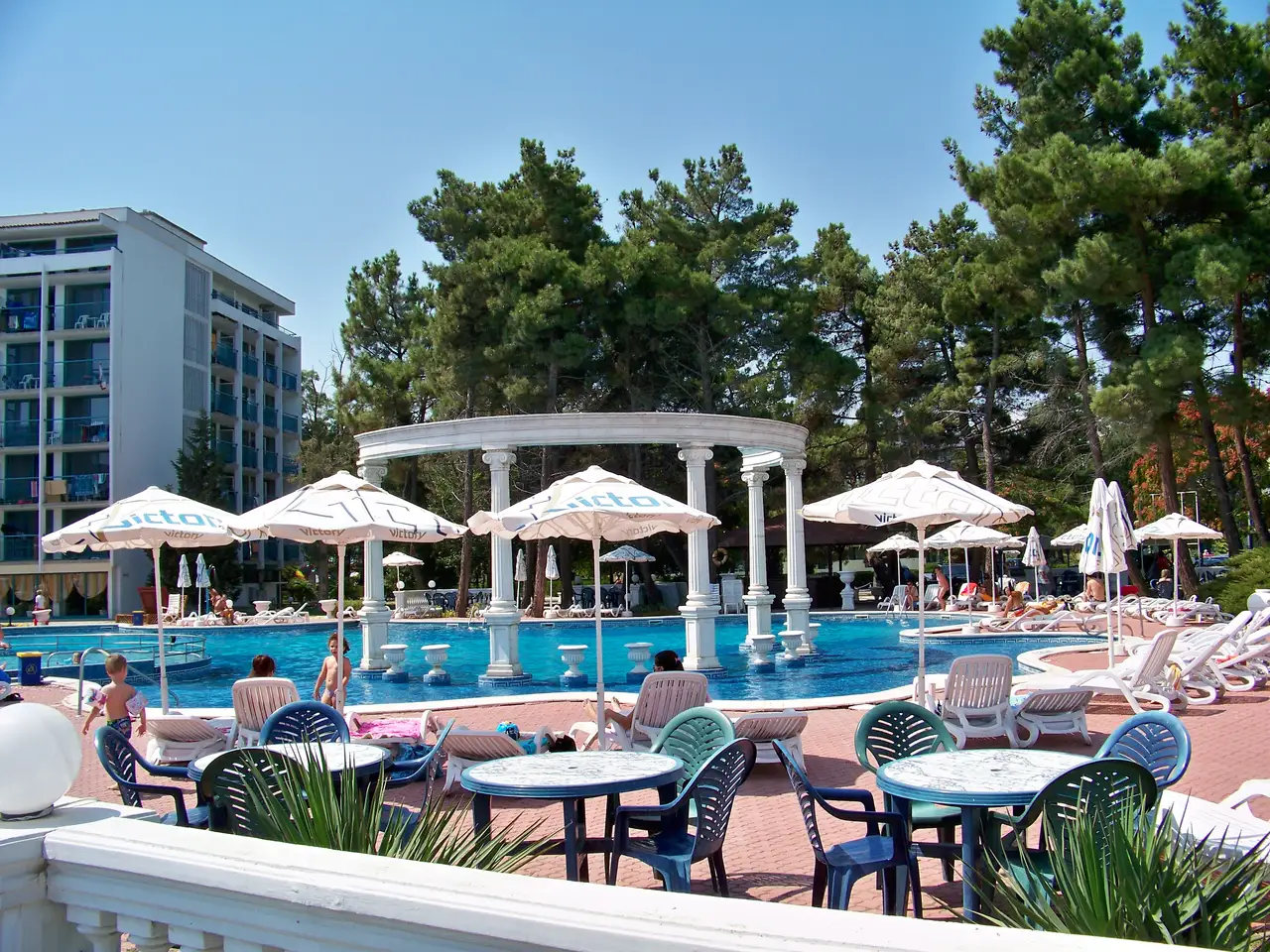 A hotel pool at Sunny Beach with a white colonnade folly in the middle, sunbeds and parasols around it and pine trees behind