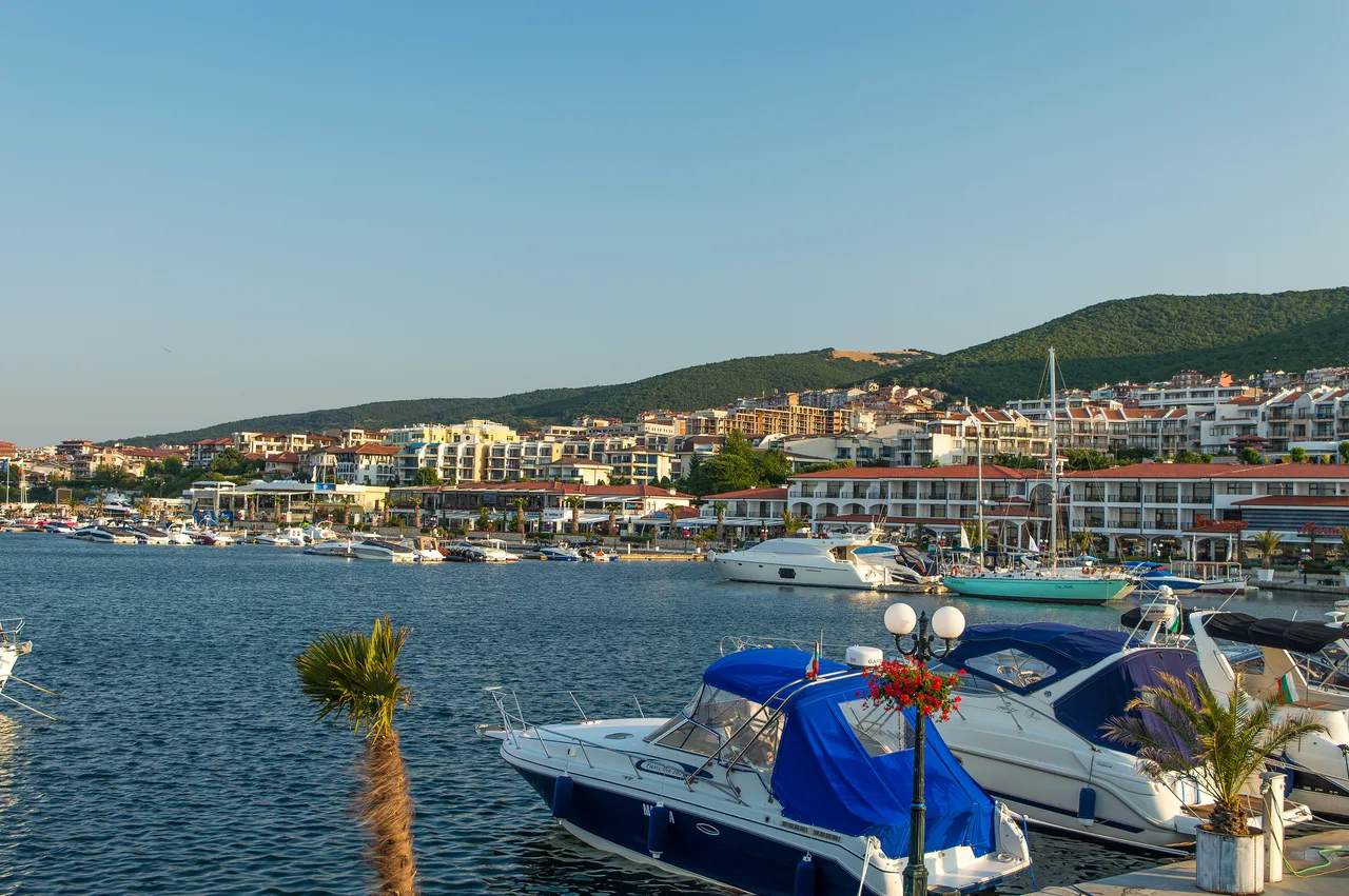 Yachts and motorboats moored at Marina Dinevi in Sveti Vlas at golden hour, low-rise white buildings and green hills rising behind the harbour