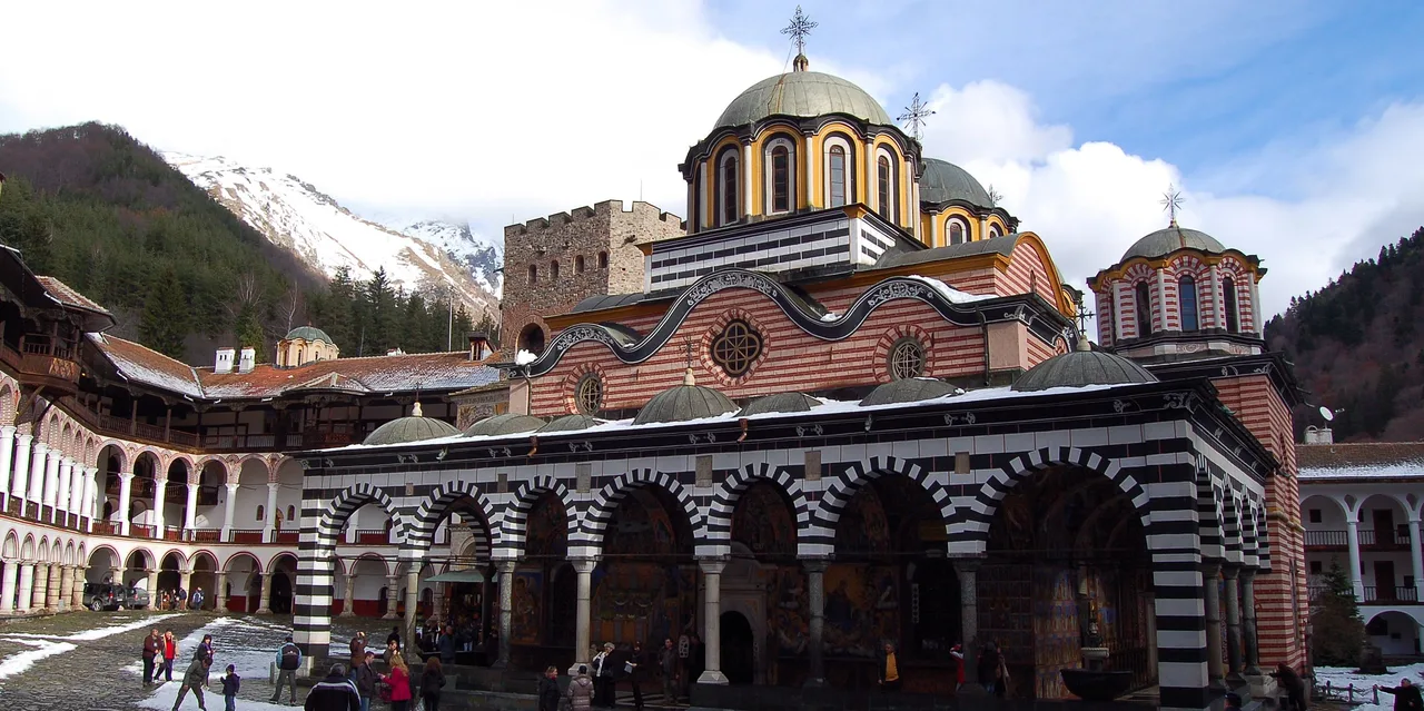 The stone courtyard of Rila Monastery with its striped arched galleries and the domed main church, mountains rising behind