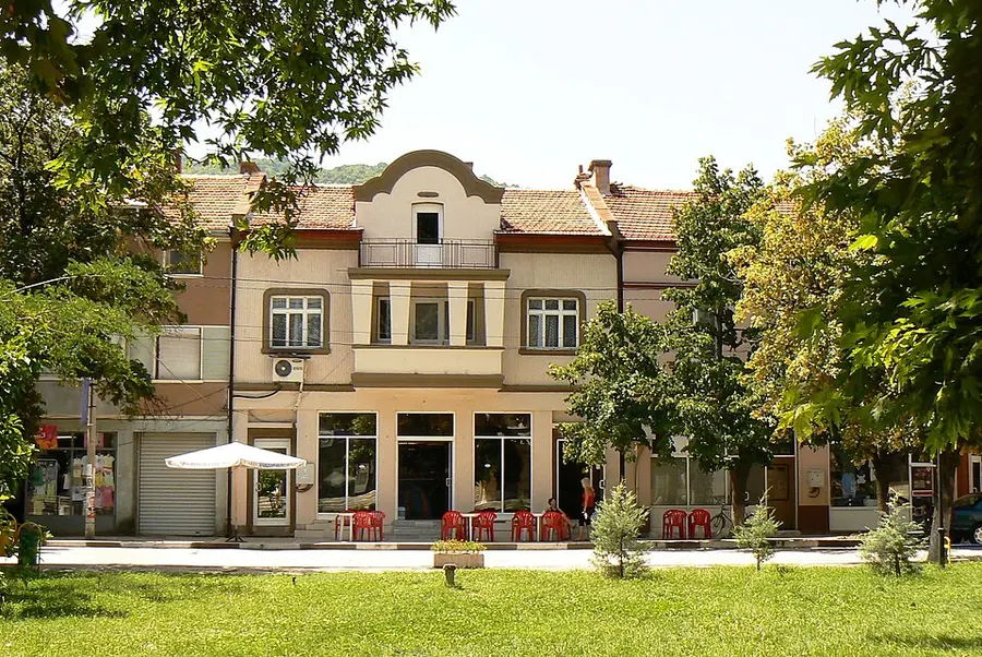 A town-square cafe in Krichim with red plastic chairs under a parasol, trees and a green park in front