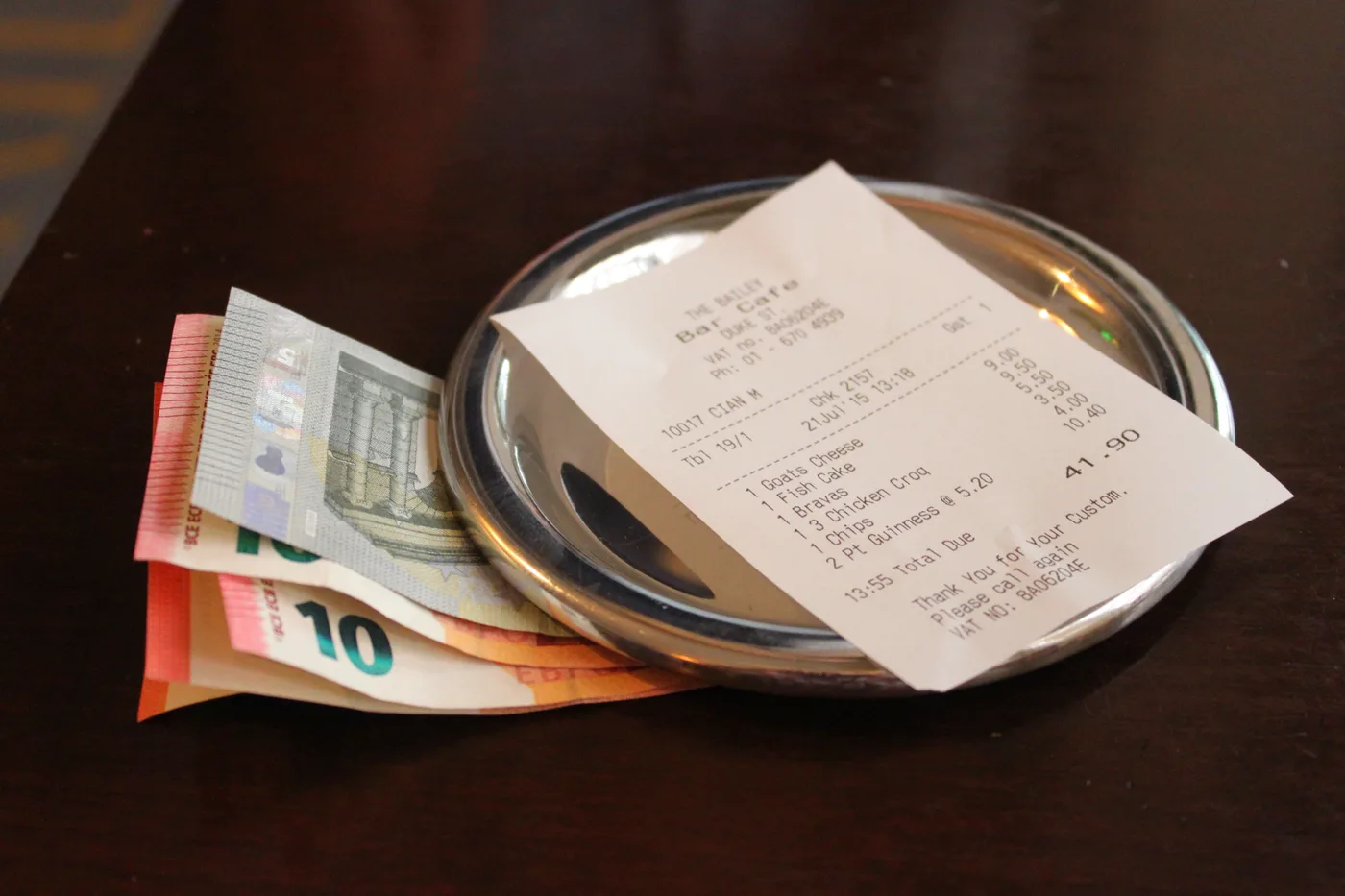 Euro banknotes tucked under a metal bill tray on a dark restaurant table, with the printed receipt resting on the tray