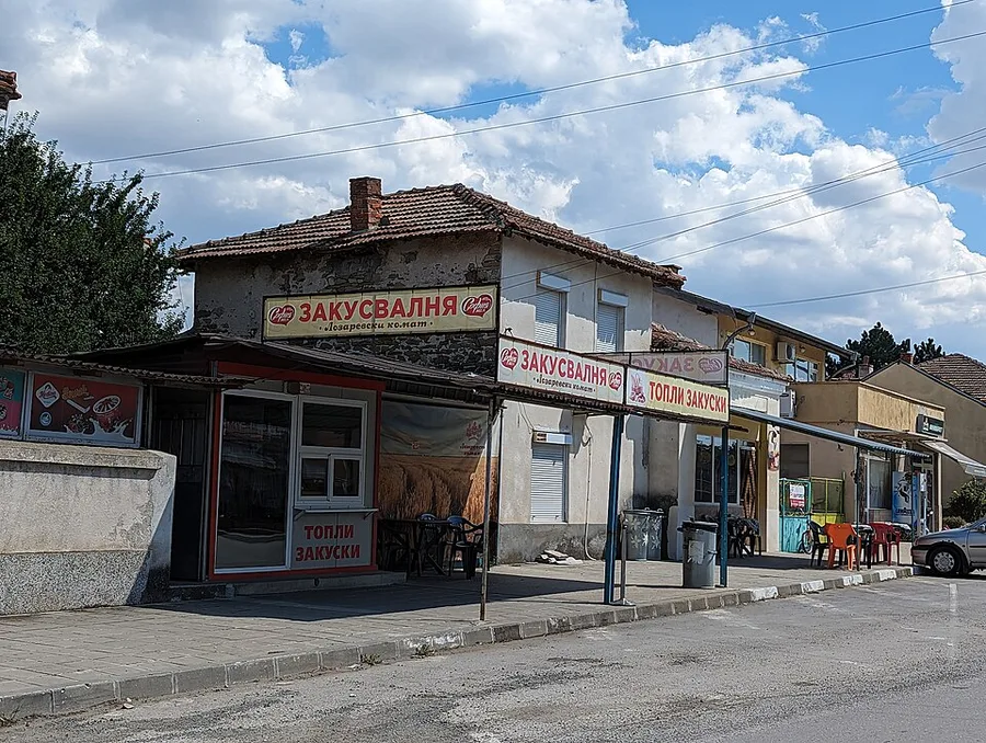 A Bulgarian snack bar with signs reading zakusvalnya and hot snacks, plastic chairs and tables outside on a small-town street