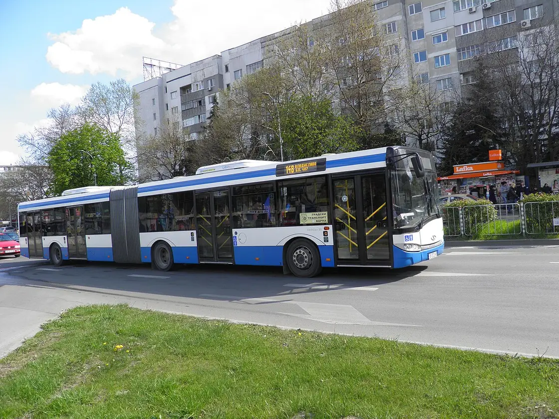 A blue and white articulated Varna city bus at a stop on a suburban street