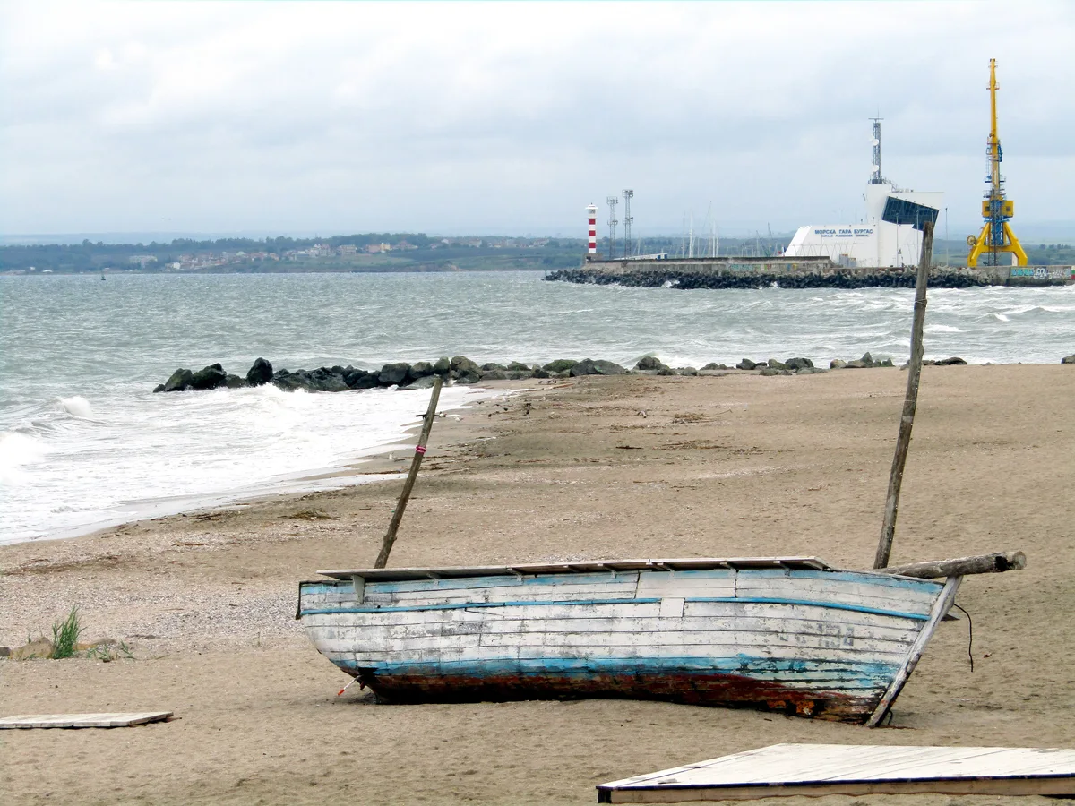 A weathered wooden fishing boat pulled up on the sand of Burgas beach, with the harbour breakwater, a yellow port crane and the sea station building across the water