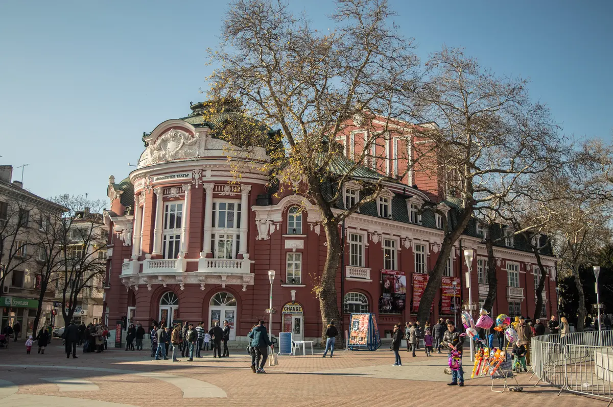 The red and white Baroque-revival facade of the Varna opera and drama theatre on a city square in winter, with bare plane trees and people crossing in coats