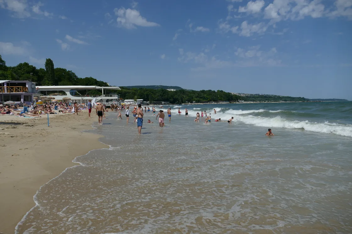 Varna's central city beach in summer: golden sand, swimmers in the shallows, beach bars on the left and a green headland behind