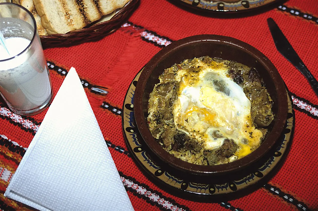 A clay pot of kavarma, a Bulgarian meat stew topped with a baked egg, on a red woven mat with bread and a glass of ayran