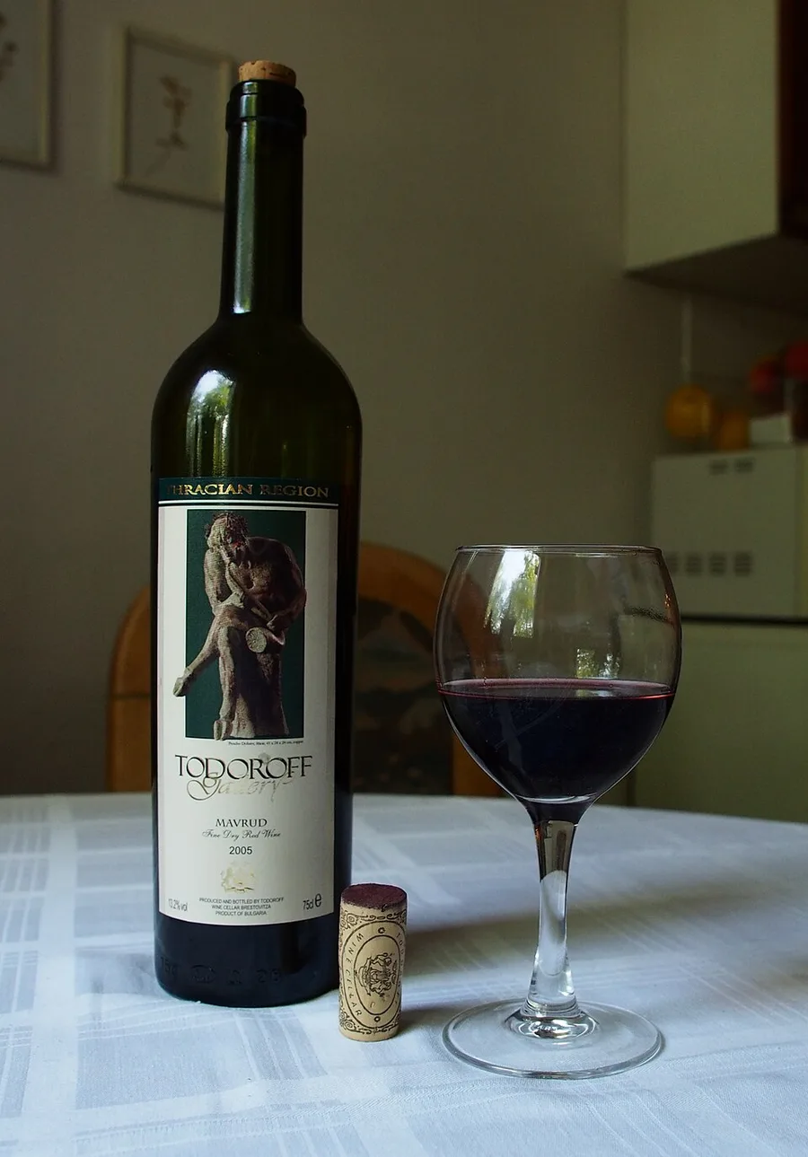 A bottle of Bulgarian Mavrud red wine from the Thracian region beside a glass of red wine on a table