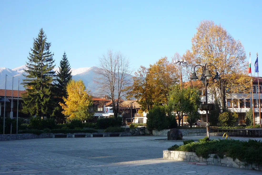 An open square in Bansko lined with autumn trees and low houses, with the Pirin mountains rising beyond the rooftops