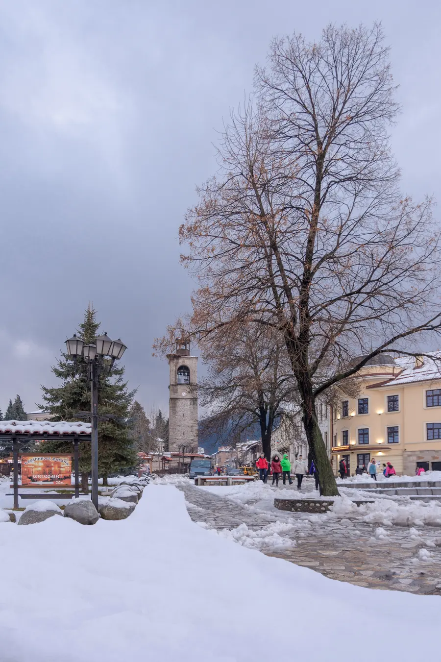 The snowy central square of Bansko with the clock tower of Holy Trinity church rising behind bare trees